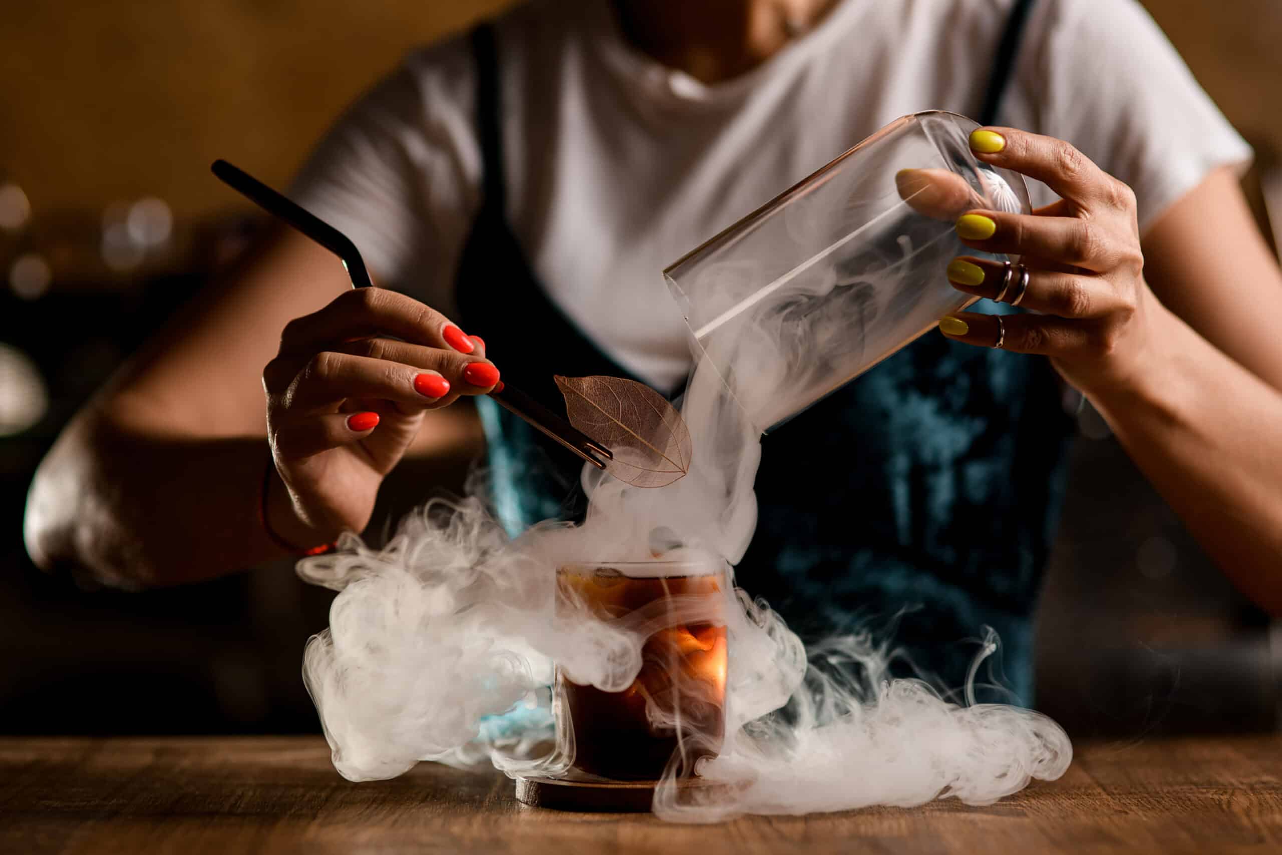 view of glass with cold cocktail with wonderful white smoke around. Woman bartender decorates it with leaf of plant. Dry ice effect.